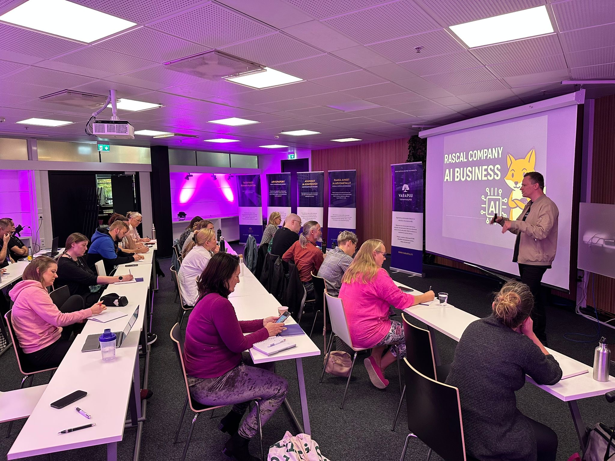 Ihmisiä istumassa luokassa riviin sijoitetuilla pöydillä, kuuntelemassa esitystä projektorin heijastaman 'Rascal Company AI Business' -kalvon äärellä.